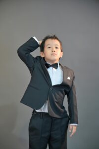 A young boy confidently poses in a classic black tuxedo with a bow tie, suitable for formal events or fashion themes.