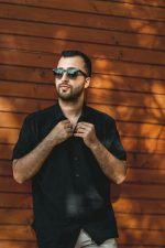 Home A fashionable man poses in sunglasses against a wooden wall, showcasing modern style.