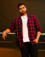 Home Portrait of a stylish young man with a beard leaning on a railing, wearing a casual plaid shirt outdoors.